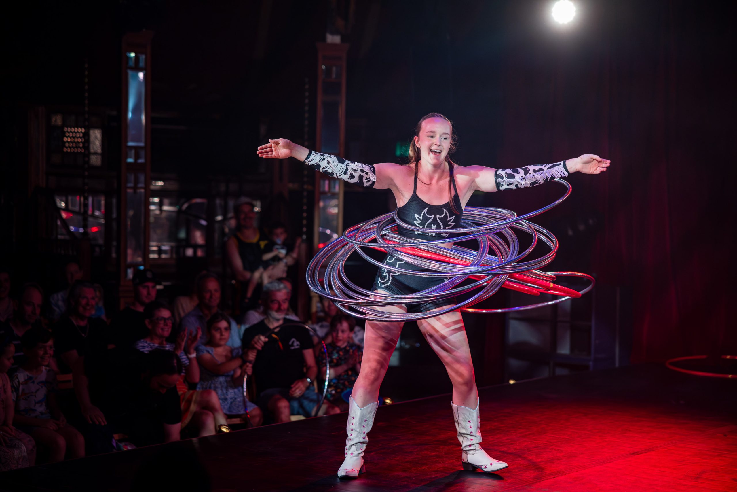 Chelsea Quaill spinning silver hula hoops on a cabaret stage in white cowboy boots